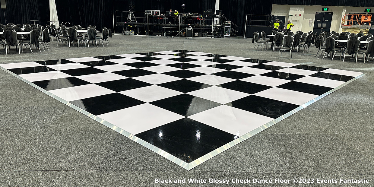 A large black and white shiny checkered dance floor set up in a hall with chairs arranged around it.